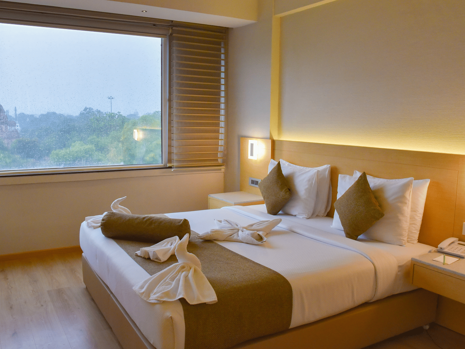 Modern hotel room at Gemini Continental featuring a neatly made double bed with accent lighting, towel art, bedside tables, and a large window offering a scenic view of greenery and a distant monument.